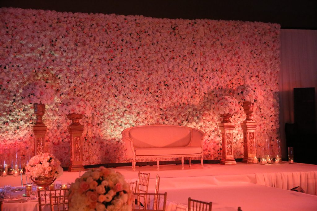 Elegant wedding stage with a vintage beige sofa against a lush pink floral wall. Ornate urns with roses flank the sofa. Soft, romantic ambiance.