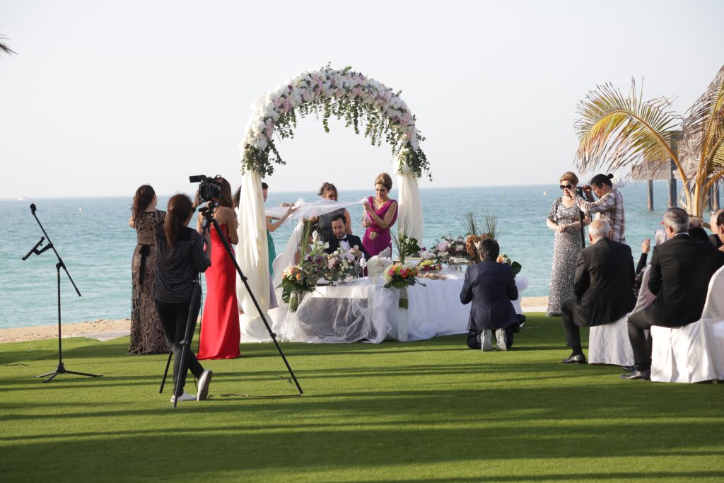 Outdoor beach wedding ceremony under a floral arch with guests seated on lush grass. A photographer captures the moment, conveying joy and celebration.