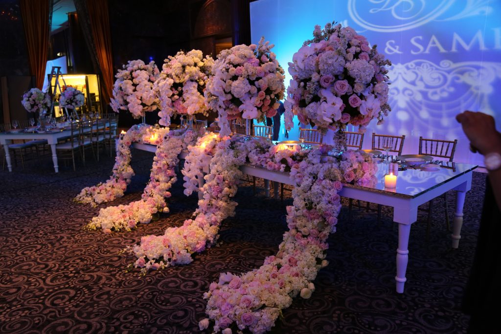 Luxurious wedding table with cascading pink and white floral arrangements, reflected in a mirrored surface. Soft candles and elegant chairs set a romantic tone.