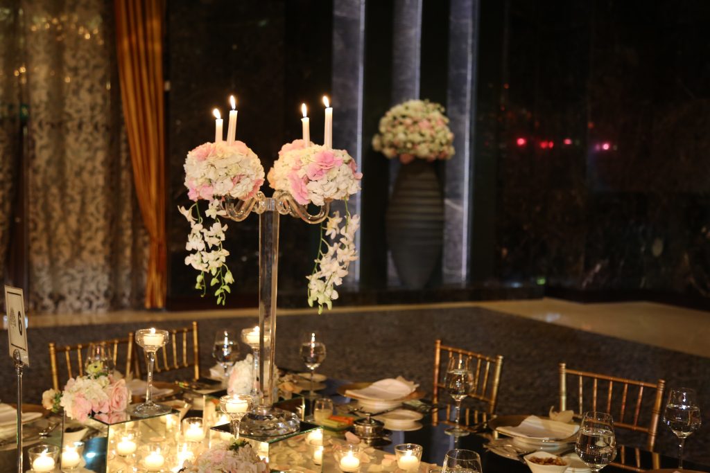 Elegant dining setup with a tall candelabra adorned with pink and white flowers. Soft candlelight and a vase of flowers create a warm, romantic ambiance.