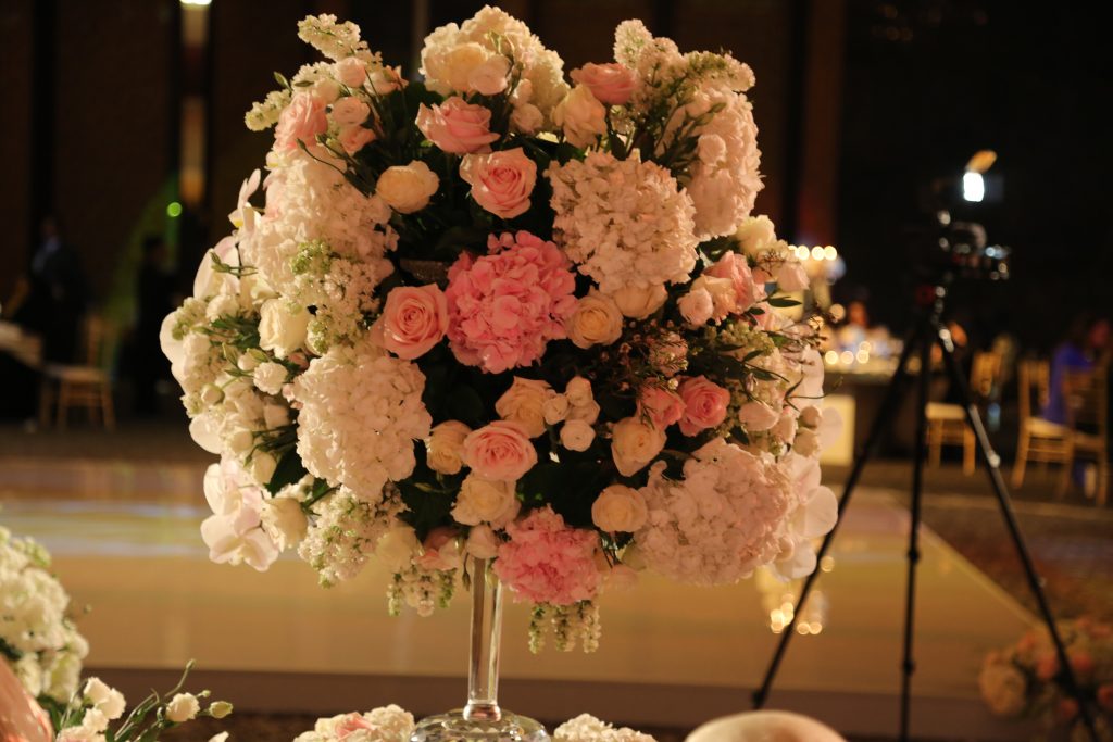 Elegant floral arrangement with soft pink and white roses on a tall stand, set in a warmly lit, sophisticated event venue.