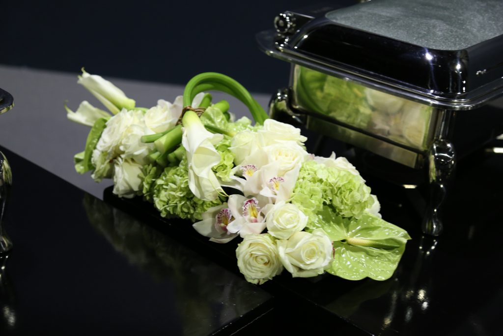Elegant white and green floral arrangement with roses, calla lilies, and orchids on a glossy black surface beside a stainless steel chafing dish.