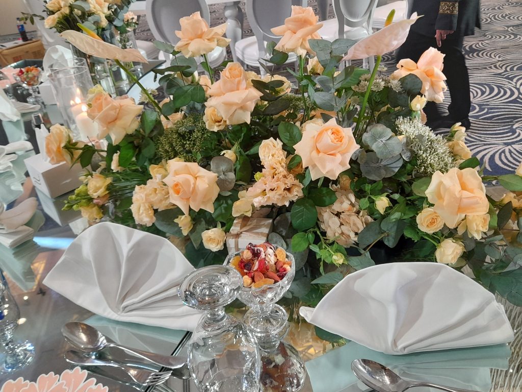 Elegant table setting with a lush bouquet of peach roses and greenery. Napkins are neatly folded, and crystal glasses sparkle alongside a small dish of dried fruit.
