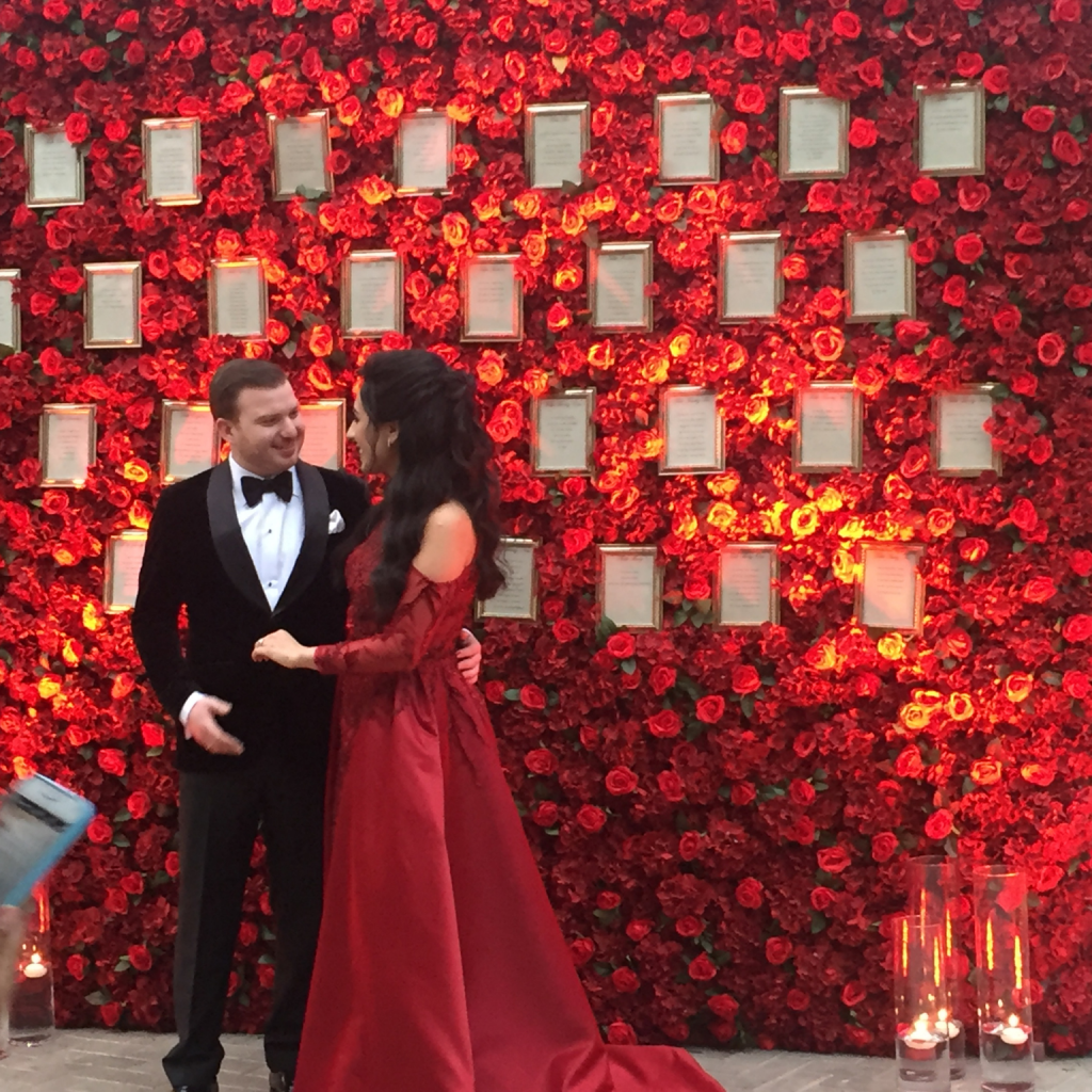 A man and woman in formal attire pose in front of a vibrant wall of red roses, showcasing a unique wedding flower theme.