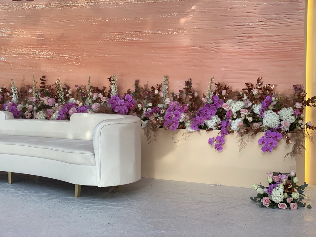 Elegant pink-themed backdrop with abundant flowers, including purple orchids and white blooms, alongside a sleek white sofa. The scene is serene and sophisticated.