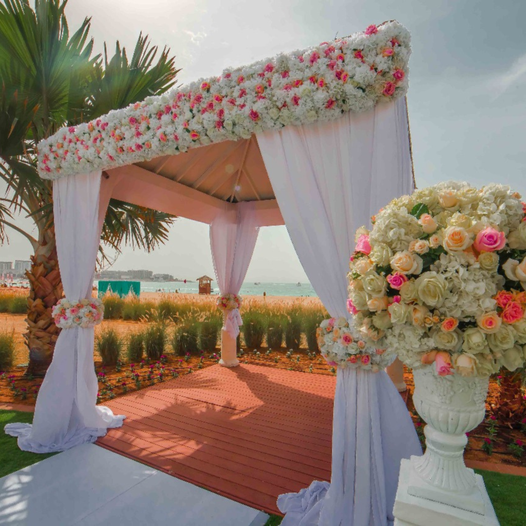 A beachside wedding arbor with white drapes and pink flowers stands under a palm tree. A bouquet of roses and peonies adds romance to the sunny setting.