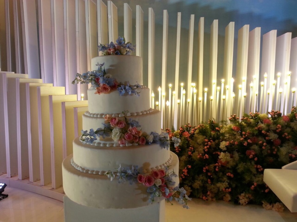 Elegant four-tier white wedding cake adorned with pastel flowers, set against a backdrop of vertical white panels and warm candlelight.