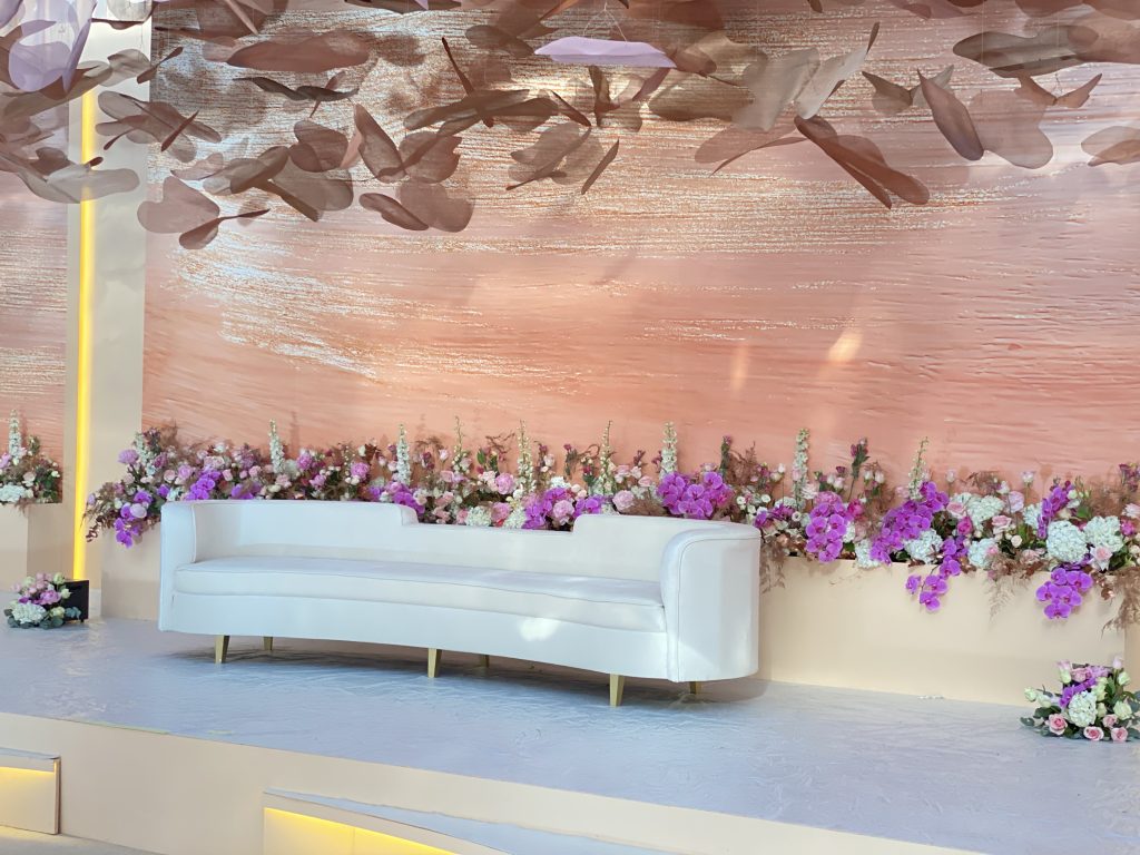 A white couch positioned in front of a floral wall, showcasing elegant decor for an Arabic wedding celebration.