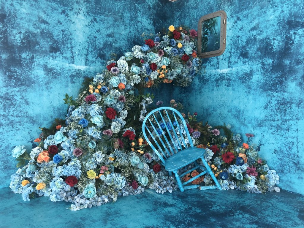 Vibrant blue room with a rustic chair and a wave of colorful flowers, including red, orange, and blue blooms. A small window reflects a floral scene.
