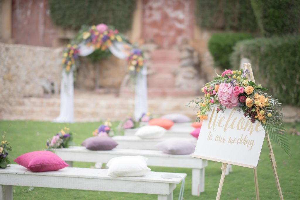 Outdoor wedding setup with a sign reading "Welcome to our wedding" adorned with colorful flowers. White benches and pillows lead to a floral archway. Serene and festive.