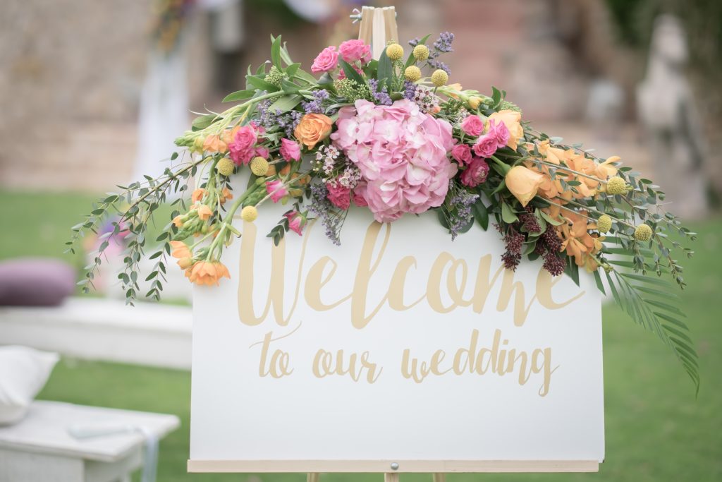 A white sign with gold script reading "Welcome to our wedding," adorned with vibrant pink, orange, and yellow flowers, set in a lush green garden.