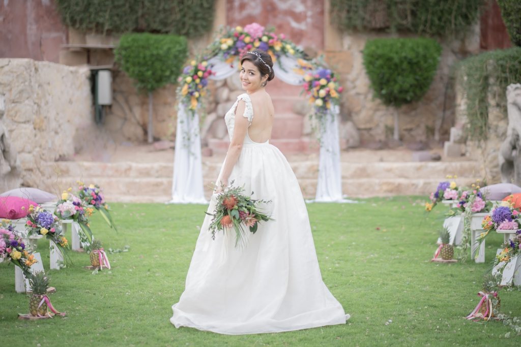 Bride in a white dress holding a bouquet, standing on a grassy aisle adorned with colorful flowers. An arch draped with flowers and fabric frames the background.