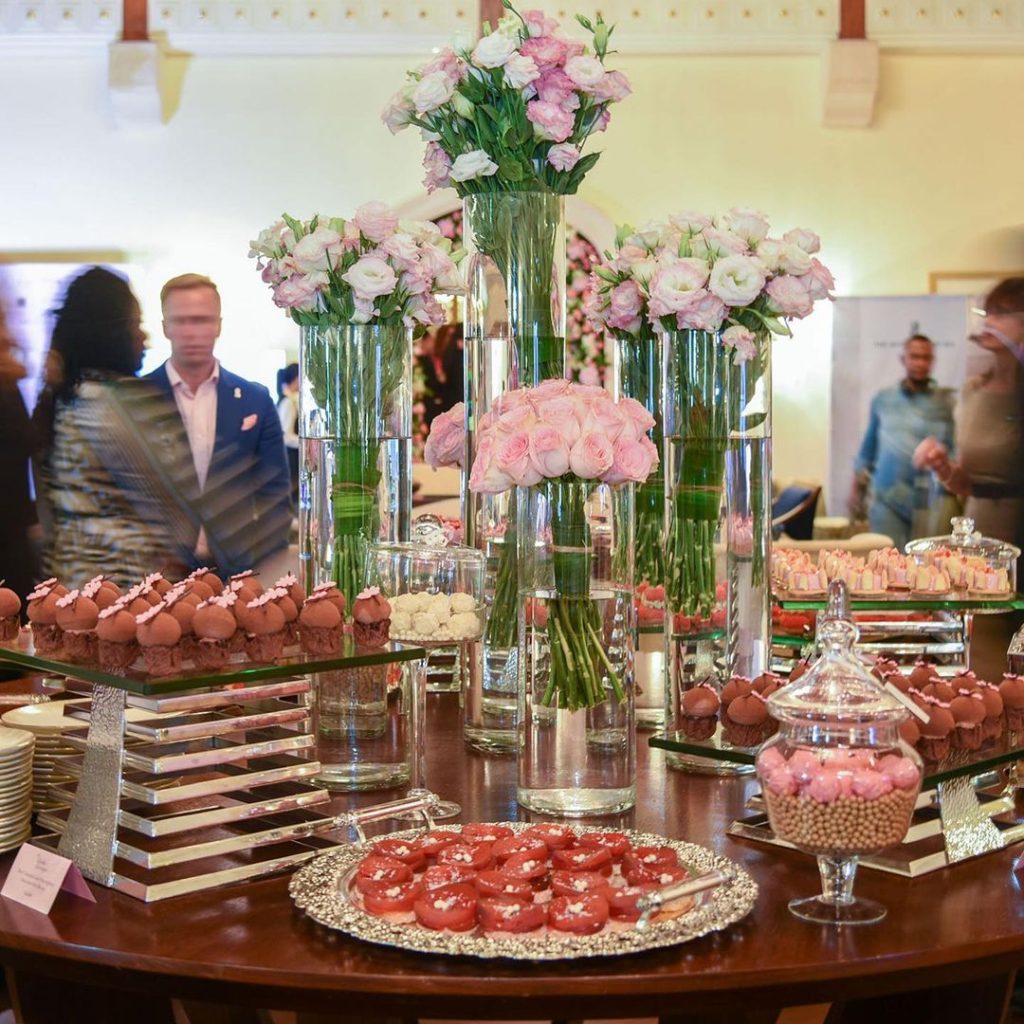 A lavish dessert display features pink and white flowers in glass vases, surrounded by delicate pastries and macarons on silver trays.