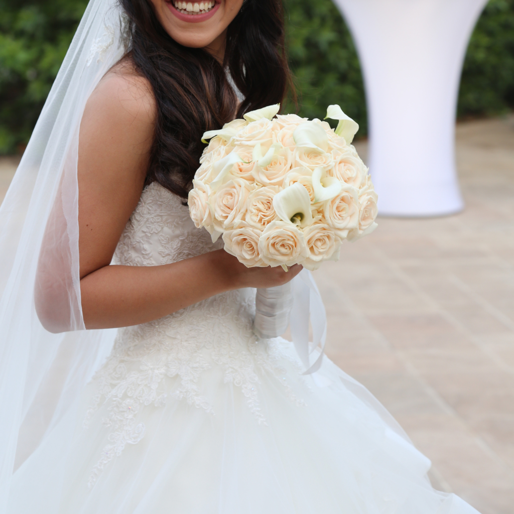 A bride in a lace wedding gown holds a bouquet of ivory roses and calla lilies, smiling warmly. The background is blurred greenery and stone tiles.