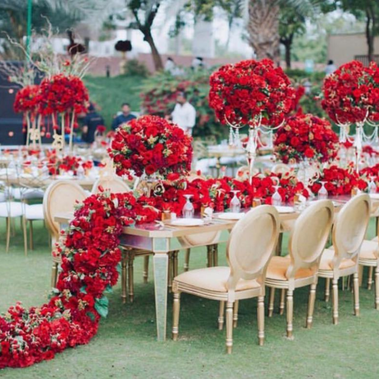 Elegant outdoor event setting with lavish red rose centerpieces and cascading floral arrangements on a gold-accented table, creating a romantic ambiance.