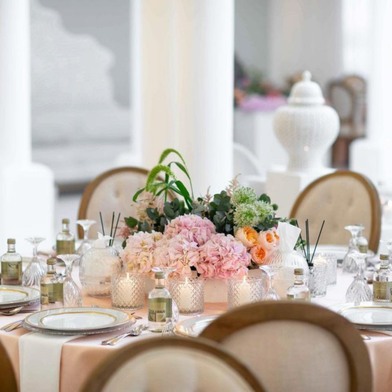 Elegant rooftop wedding table setting with pink and orange floral arrangements, crystal glassware, and soft candlelight on a peach tablecloth. City skyline in the background, creating a sophisticated open-air ambiance.
