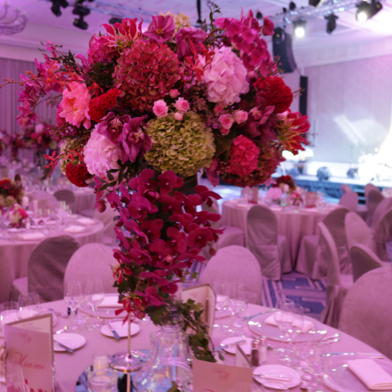 Elegant floral centerpiece with vibrant pink, red, and green flowers decorates a round table set with fine china and crystal at a formal event venue.