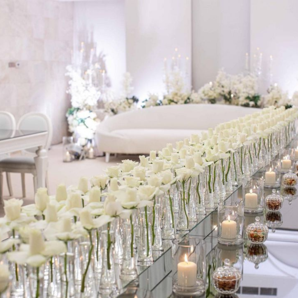 Luxurious setting with rows of white roses in glass vases on a mirrored table, surrounded by lit candles. Elegant white sofa and floral decor in the background.