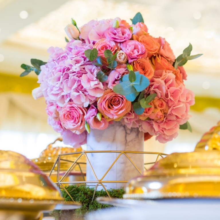 Vibrant pink roses and hydrangeas in a marble vase, surrounded by a decorative gold frame. The setting is elegant and festive, with soft, warm lighting.