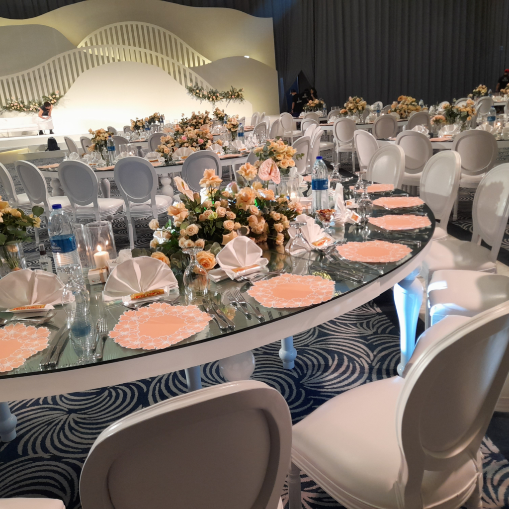 Elegant event setup with oval tables featuring peach placemats, neatly folded napkins, and floral centerpieces.