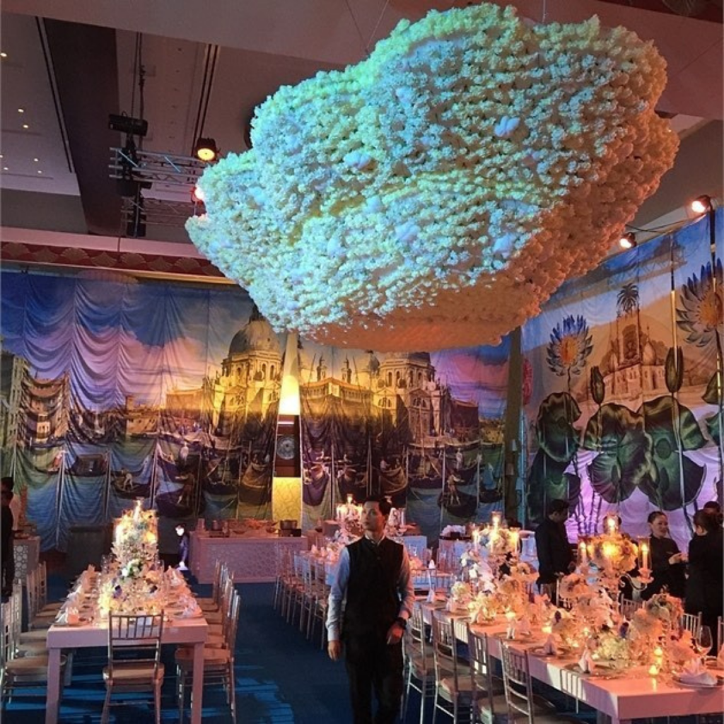 Lavishly decorated event hall with an ornate flower cloud installation and vibrant Venetian-themed murals. Tables are elegantly set, exuding a festive ambiance.
