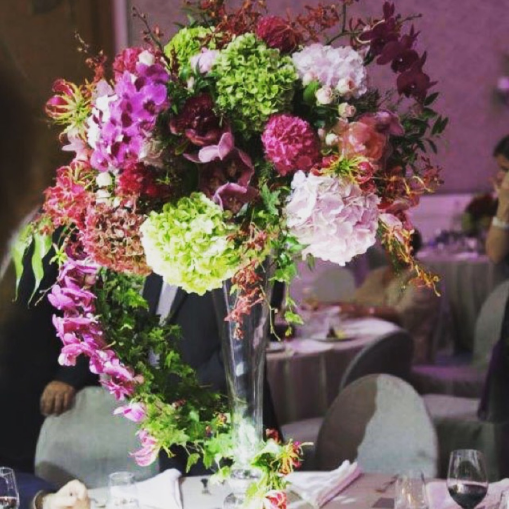 Elegant tall centerpiece with vibrant pink, purple, and green flowers, set in a clear vase on a table in a dimly lit event space, creating a luxurious feel.
