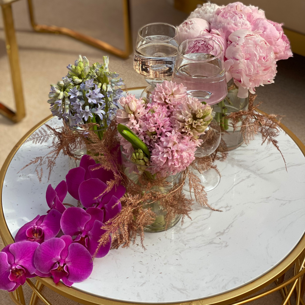 Elegant floral arrangement on a round marble table. Includes pink peonies, purple orchids, and hydrangeas in glass vases, exuding a luxurious, peaceful ambiance.