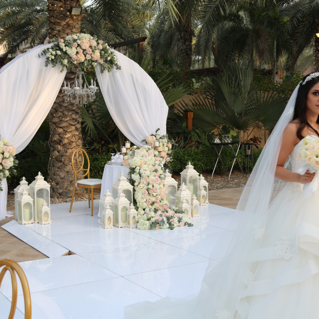 A bride in a flowing white gown holds roses, standing beside a decorated arch with sheer drapes and flowers.